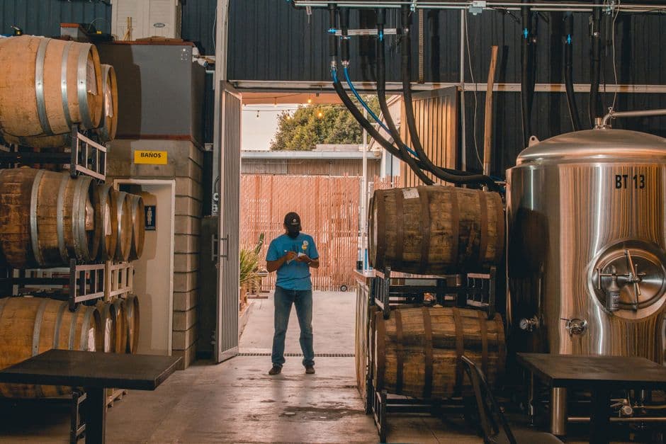 Two brewers in a dimly lit brewery examining small-batch beer samples in glass vessels, surrounded by barrels and brewing equipment, with a focused and experimental atmosphere