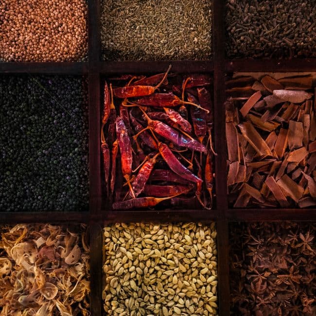 A flat-lay of unusual brewing ingredients arranged on a dark slate surface — dried hibiscus, toasted cardamom pods, charred oak chips, and a small jar of saffron — representing the global flavor inspirations behind a Flansk limited release